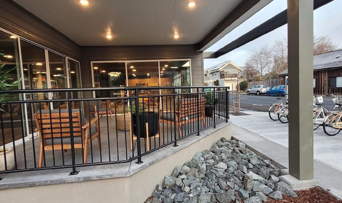 The image shows the exterior of a building labeled Sorrel Place with a modern overhang, patio seating, and a small rock pile. Bicycles are parked nearby on a concrete sidewalk, with trees and other buildings in the background.