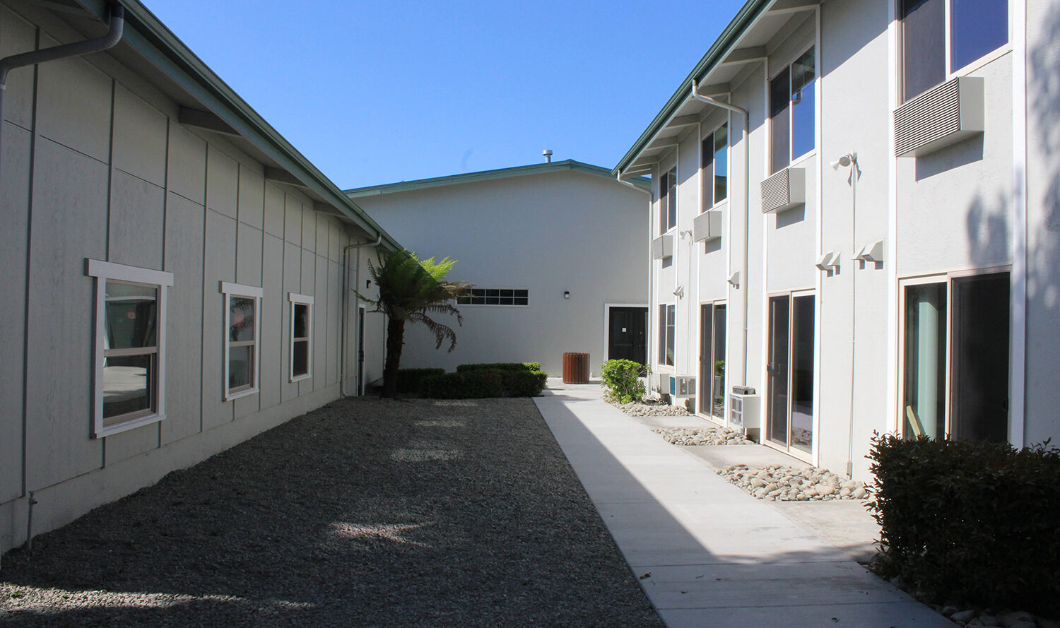 A narrow courtyard between two light-colored buildings with multiple windows, a small tree, and pebbled ground. The pathway leads to a door at the far end. The sky is clear and blue.