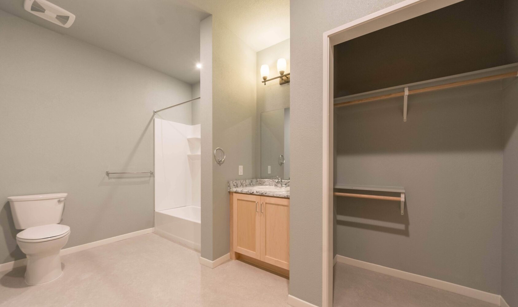 A spacious bathroom with a toilet, shower, vanity with a marble countertop, and a light fixture above the mirror. Theres an open closet with a shelf and hanging bar on the right side. The walls are painted light gray.