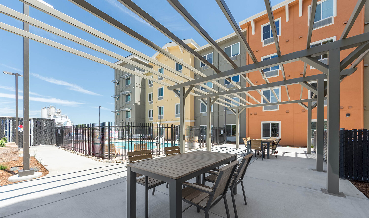Outdoor seating area under a pergola with wooden tables and chairs, near a fenced swimming pool. Background shows multi-story buildings with a mix of tan and orange exteriors under a clear blue sky.