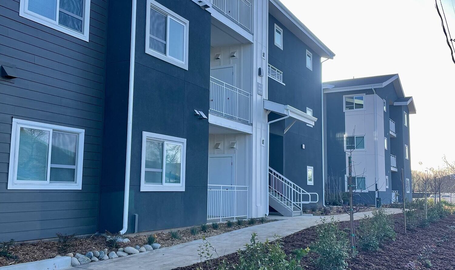 A modern apartment building with blue-gray and white accents, featuring three stories and balconies. The landscaping includes young plants and a mulch-covered area beside a concrete walkway under a clear sky.