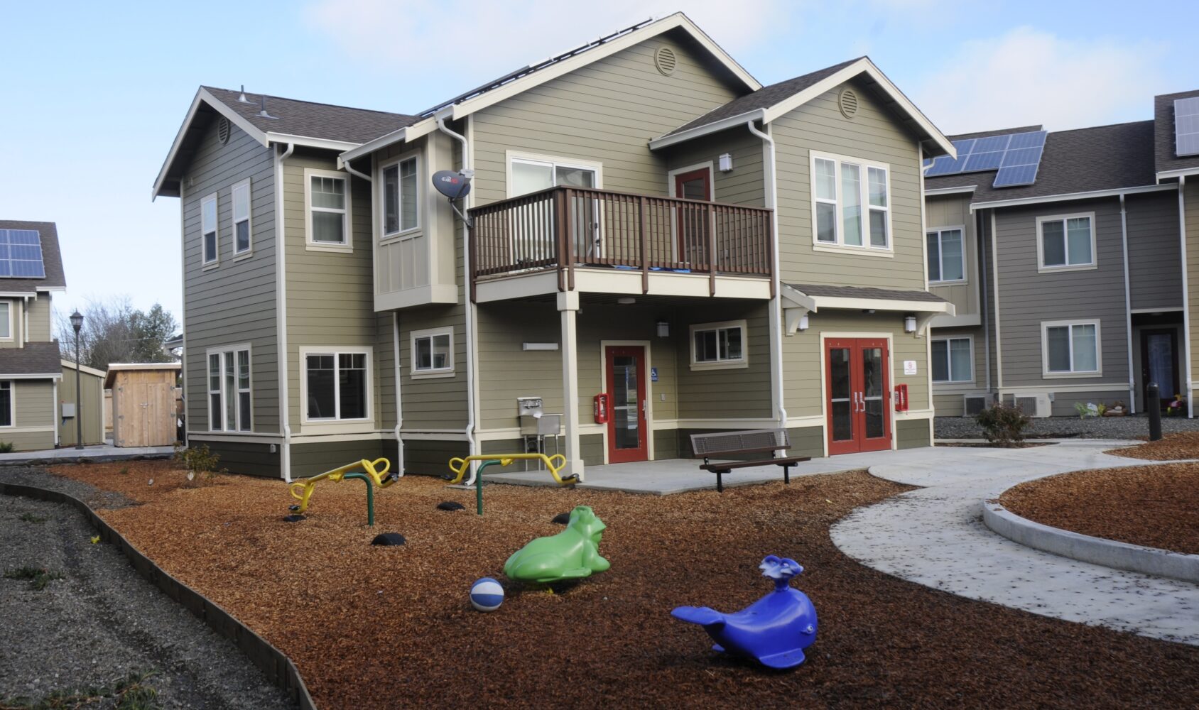 A modern two-story building with beige and green siding, featuring balconies and white trim. The yard has a playground with animal-shaped equipment and a bench. Solar panels are visible on the rooftops under a partly cloudy sky.