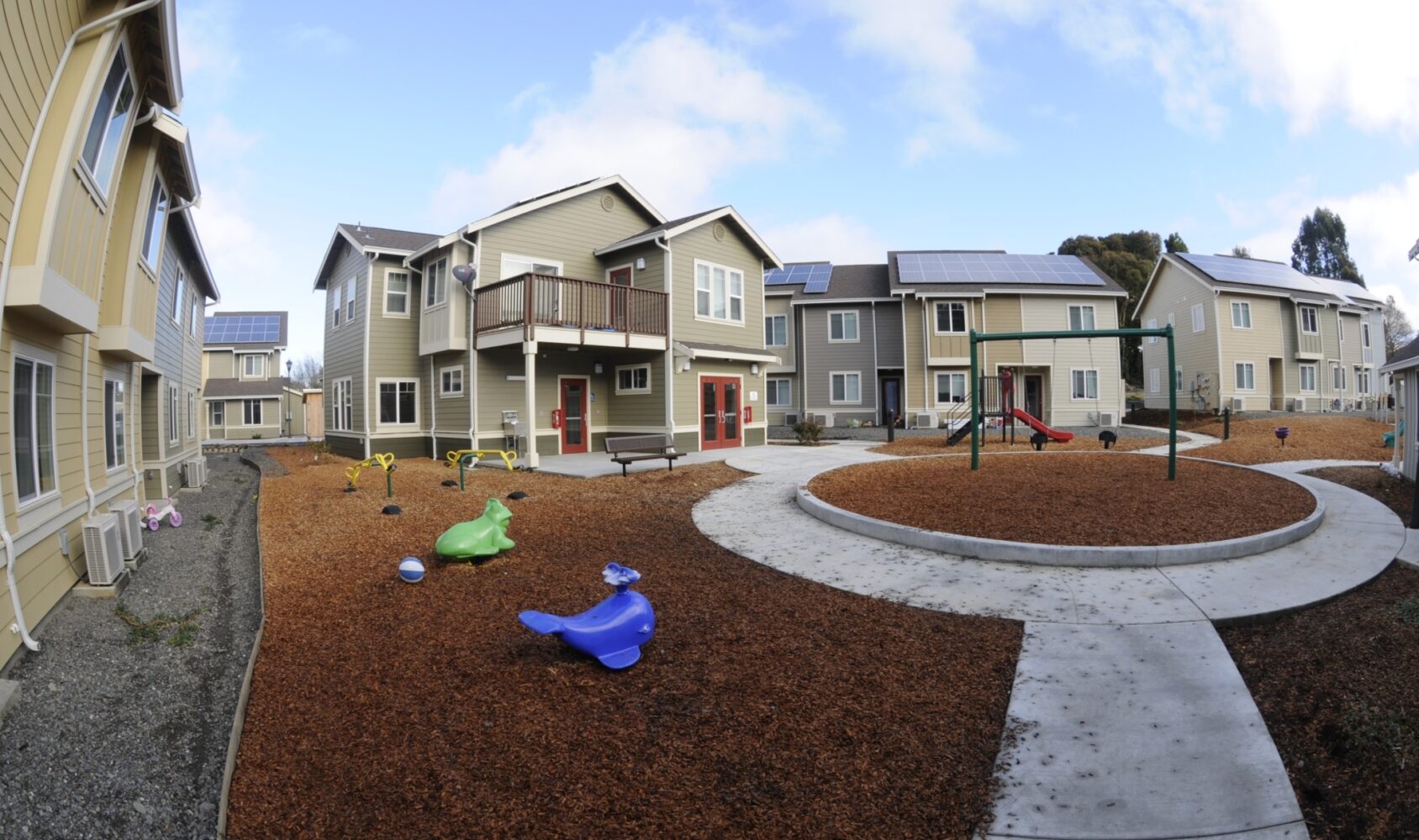 A modern apartment complex with earth-toned buildings surrounds a playground area featuring a swing set and colorful animal-shaped spring riders. Solar panels are visible on the rooftops, and the sky is partly cloudy.
