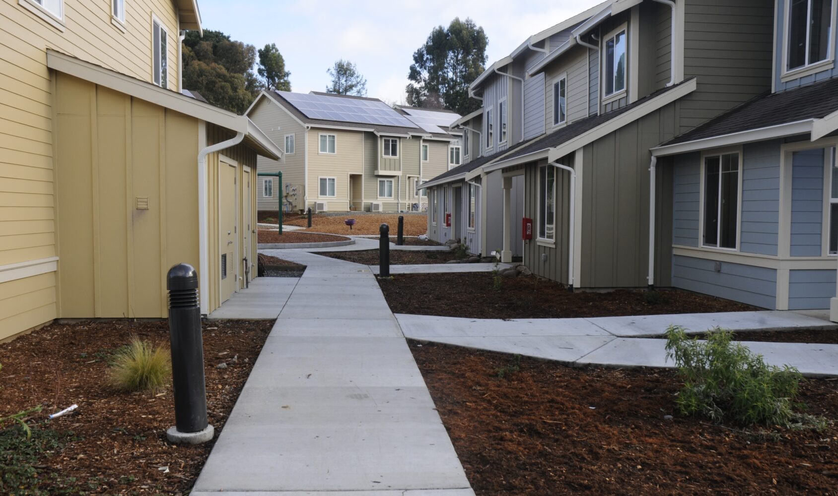 A residential area with multiple two-story beige and gray houses, a central concrete pathway, and landscaped grounds. Solar panels are visible on one house. The scene is set on a clear day with trees in the background.