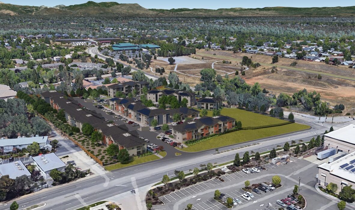 Aerial view of a suburban area with residential buildings, roads, and greenery. The foreground features a parking lot and a street with vehicles, while the background shows hills and a mix of trees and houses.