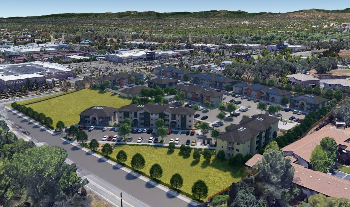 Aerial view of a suburban area featuring a complex of residential buildings surrounded by green spaces. Nearby, there are commercial buildings and parking lots. In the background, there are hills and a mix of trees and roads.