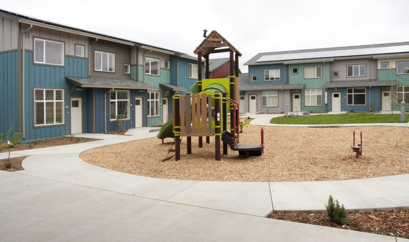 A small playground with a climbing structure and slide is in the center of a courtyard surrounded by modern two-story buildings with blue and gray exteriors. The area is landscaped with grass and mulch paths.