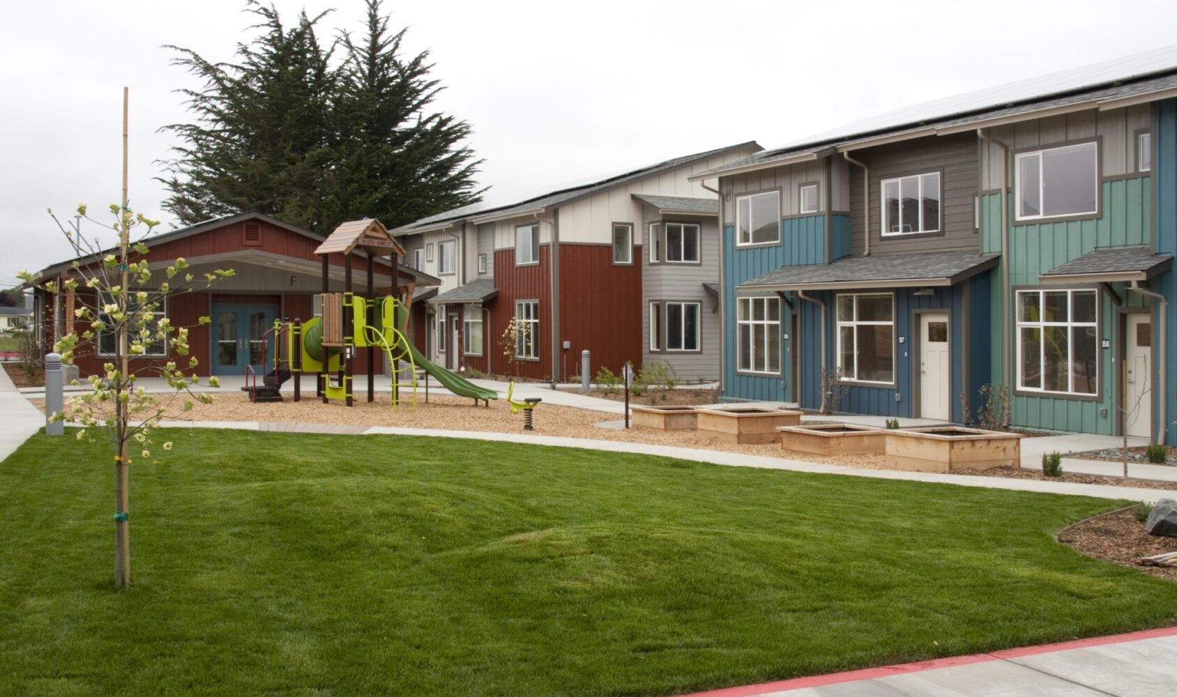 A suburban residential area with a row of modern, two-story houses in various colors. In the foreground, there is a grassy lawn, young trees, and a playground with a slide and climbing structure. Cloudy sky overhead.