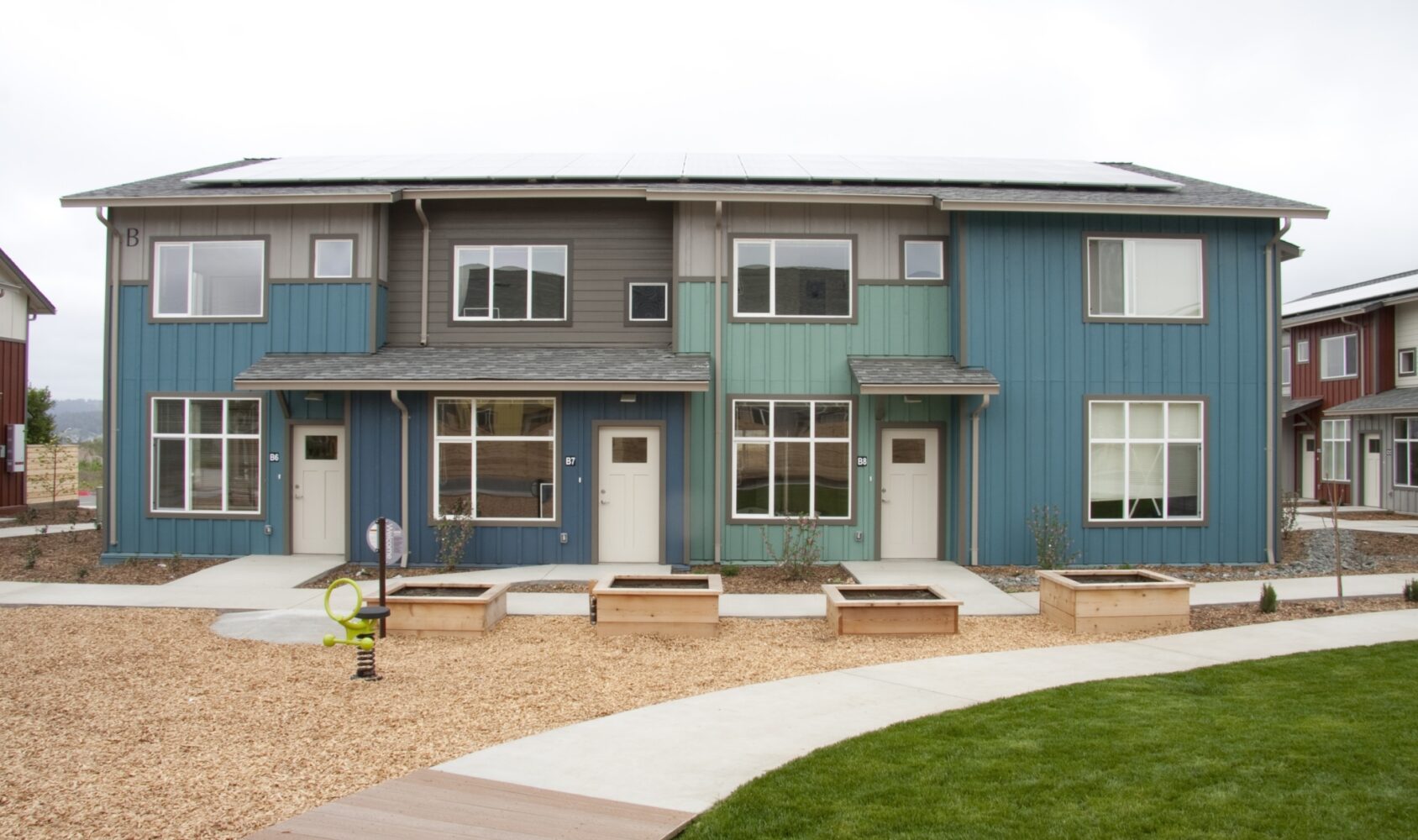 A modern, two-story building with blue and green exterior panels. It has two front doors and large windows. In front, there are wooden planters and a landscaped area with a curved pathway and a small play structure.