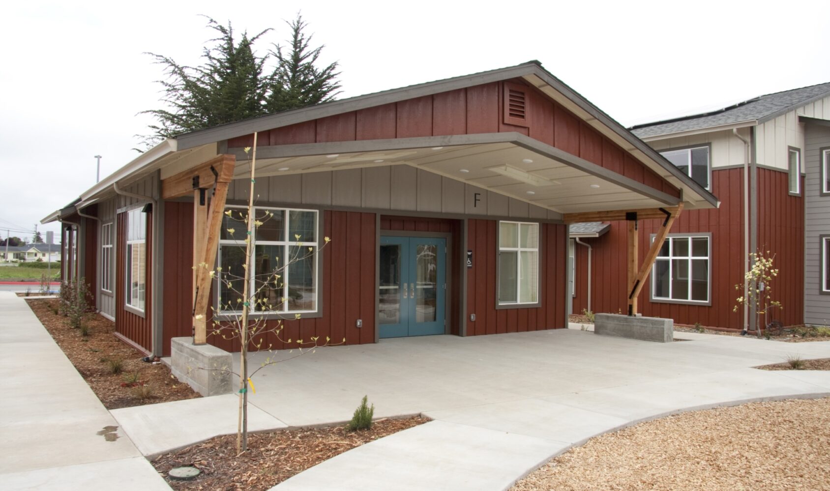 A modern wooden building with a covered entrance and blue double doors. The structure is surrounded by a landscaped area with young trees and paved walkways. The exterior features a mix of red and gray panels under a cloudy sky.
