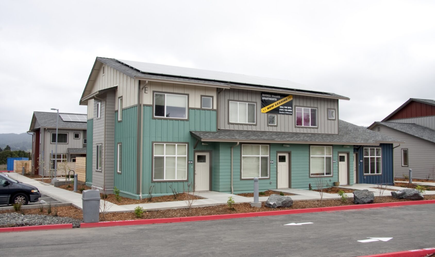 A modern two-story house with a green and gray facade, solar panels on the roof, and a Model Home sign displayed. The surrounding area includes paved pathways, rocks, and a red curb in a residential neighborhood.