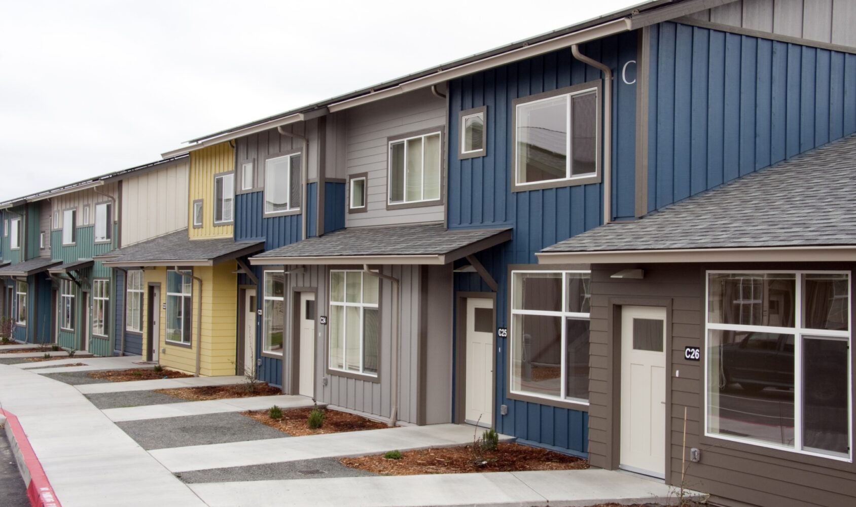 A row of modern townhouses with colorful facades in shades of blue, yellow, and gray. Each house has a small front yard with young plants and a paved walkway. The sky is overcast.