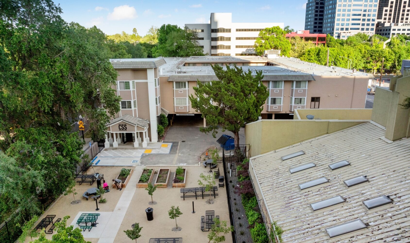 Aerial view of a building with a courtyard featuring benches, tables, and small trees. A few people are sitting and walking in the outdoor area. The background shows a mix of trees and high-rise buildings under a blue sky.