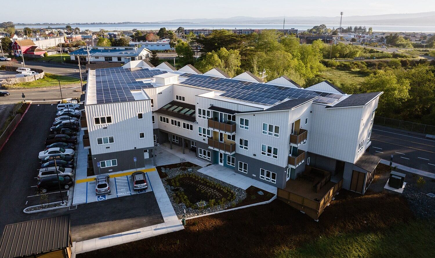 Aerial view of a modern, multi-story building with solar panels on the roof, surrounded by trees and a parking lot. Roads and other buildings are visible, with a body of water in the background under a clear sky.