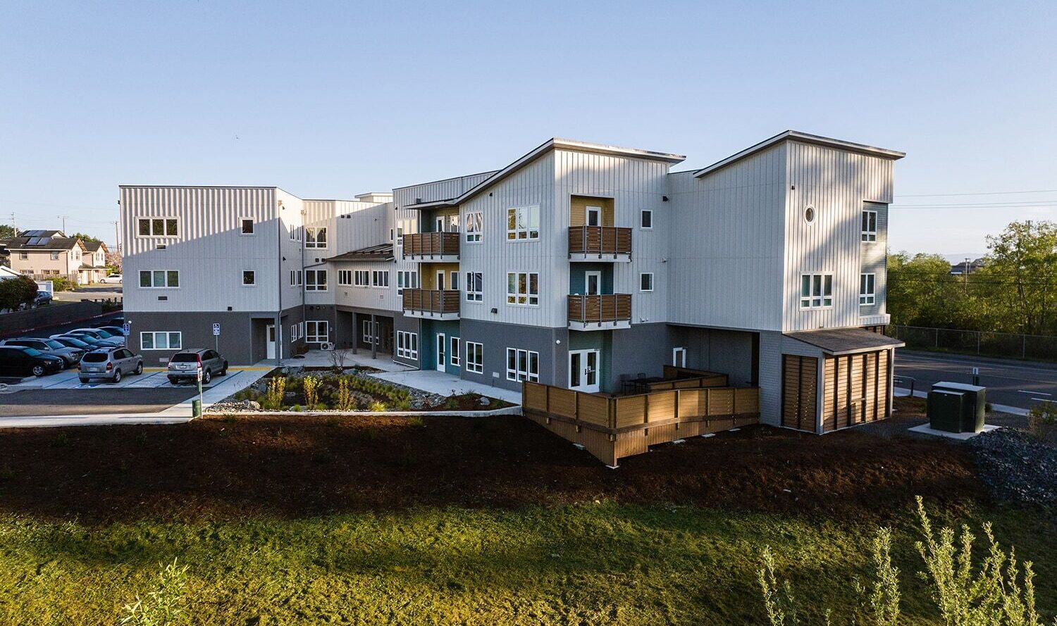 A modern apartment building with a mix of gray and white panels, featuring multiple balconies and large windows. It is surrounded by landscaped greenery and has a parking area on the left side. The sky is clear and blue.