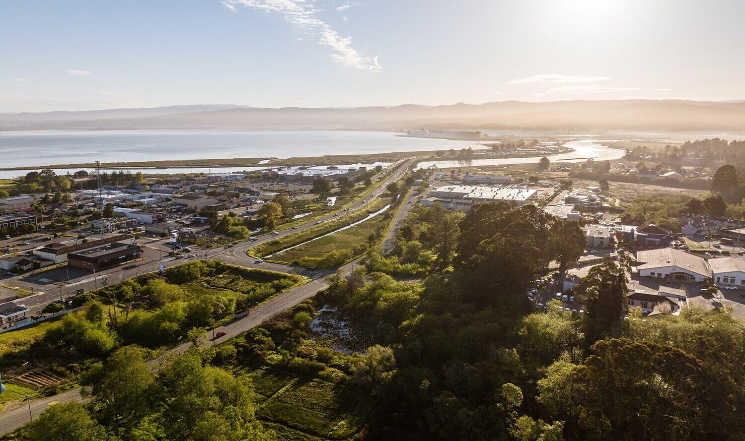 Aerial view of a coastal town with buildings and roads surrounded by lush greenery. A river winds through the landscape, and the sun shines brightly over the distant mountains and sea.