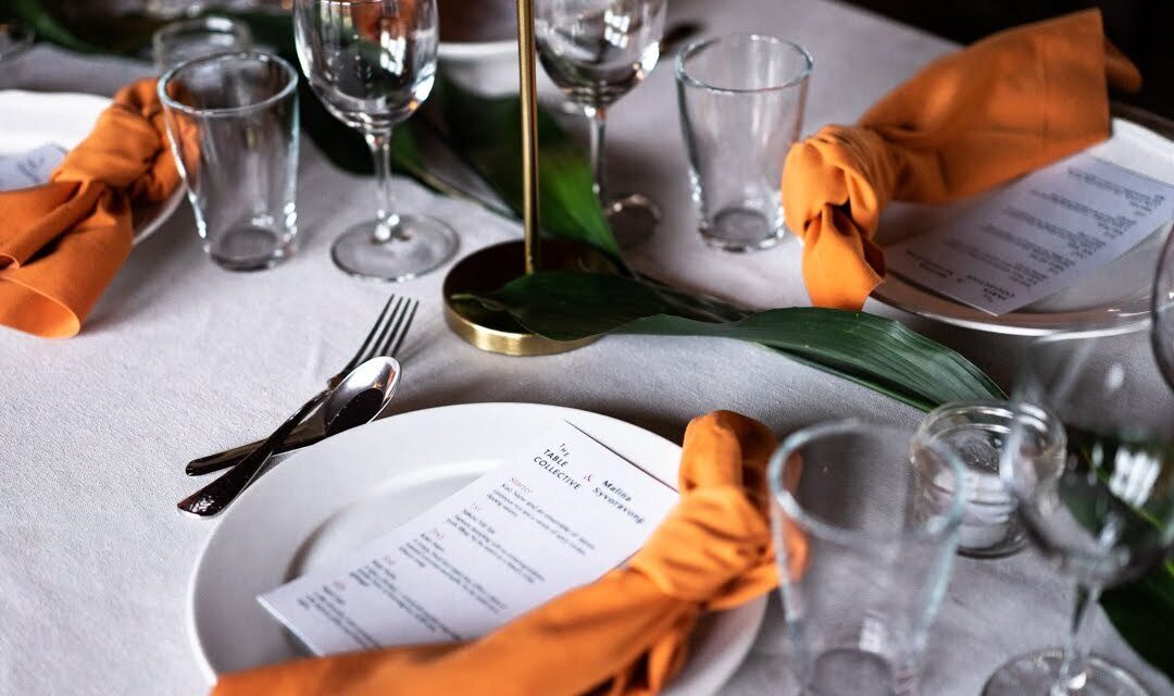 A table is elegantly set for a meal with white plates, glasses, and cutlery. Orange cloth napkins are artfully tied and placed on the plates, alongside menus. Green leaves decorate the center of the white tablecloth.
