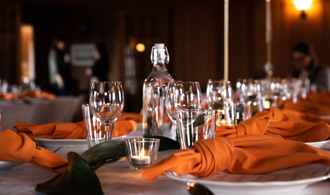 A beautifully set dining table features orange napkins, clear glasses, and a glass bottle. A small candle is lit in the foreground, and the background shows blurred figures in a warmly lit room.