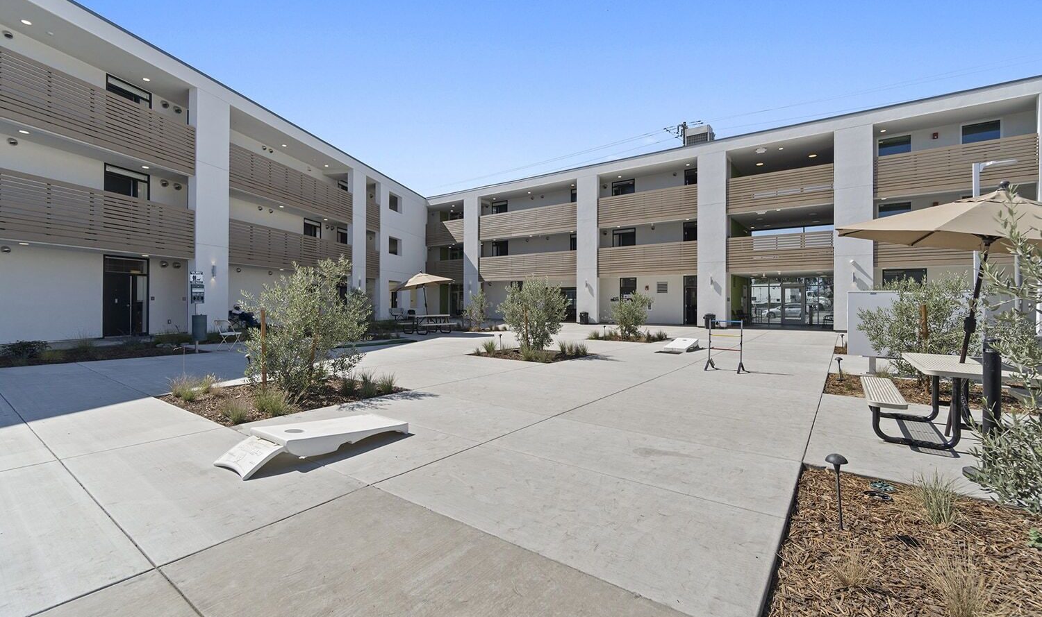 A modern apartment complex with three floors and a central courtyard. The courtyard features benches, small plants, cornhole boards, and an umbrella-covered picnic table. The building has wooden railings and a clear blue sky overhead.