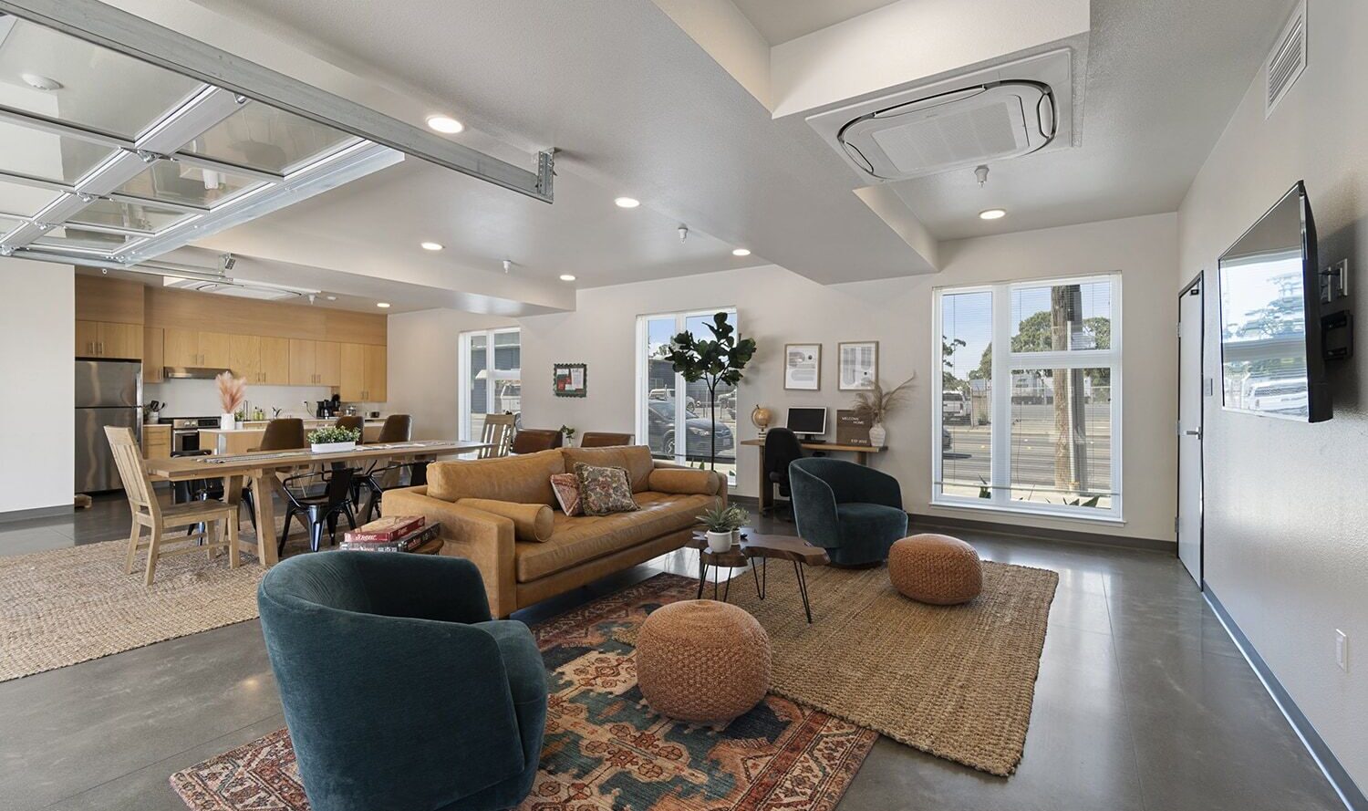 A modern, open-concept living room with a green sofa, two blue chairs, and a wooden dining table. The kitchen is in the background. Large windows bring in natural light. Rugs and plants add warmth to the space. A flat-screen TV is mounted on the wall.