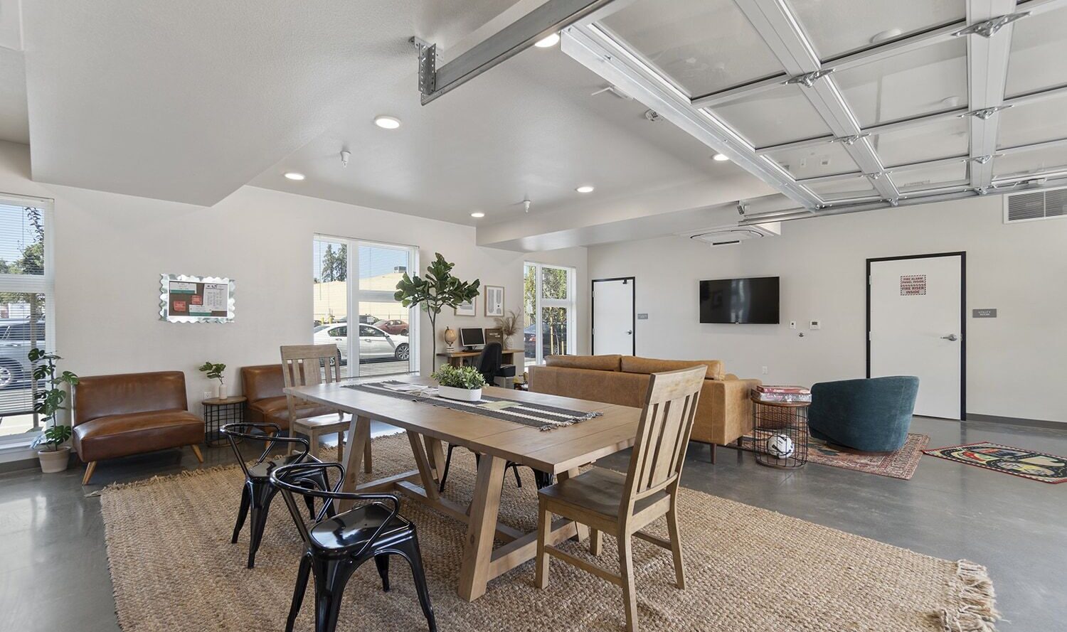 Spacious, modern living area with a light wood dining table and black chairs on a woven rug. Cozy seating area includes a brown sofa and green chair. Large windows and a glass garage-style door let in natural light. White walls and minimal decor.