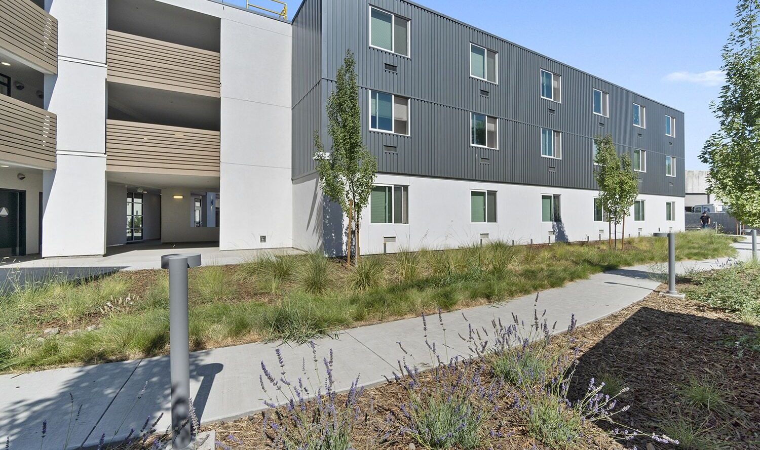 A modern apartment building with three floors featuring a combination of white and gray exteriors. The landscaped area includes grasses and small shrubs, bordered by paved walkways. The sky is clear and blue.