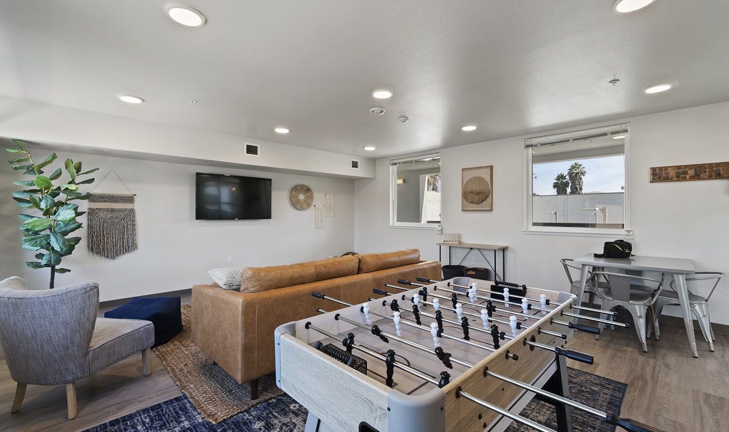 A modern lounge with a foosball table in the foreground, a brown couch, armchair, and a wall-mounted TV. Brightly lit room with a patterned rug, art pieces on walls, and a small dining area. Large windows bring in natural light.