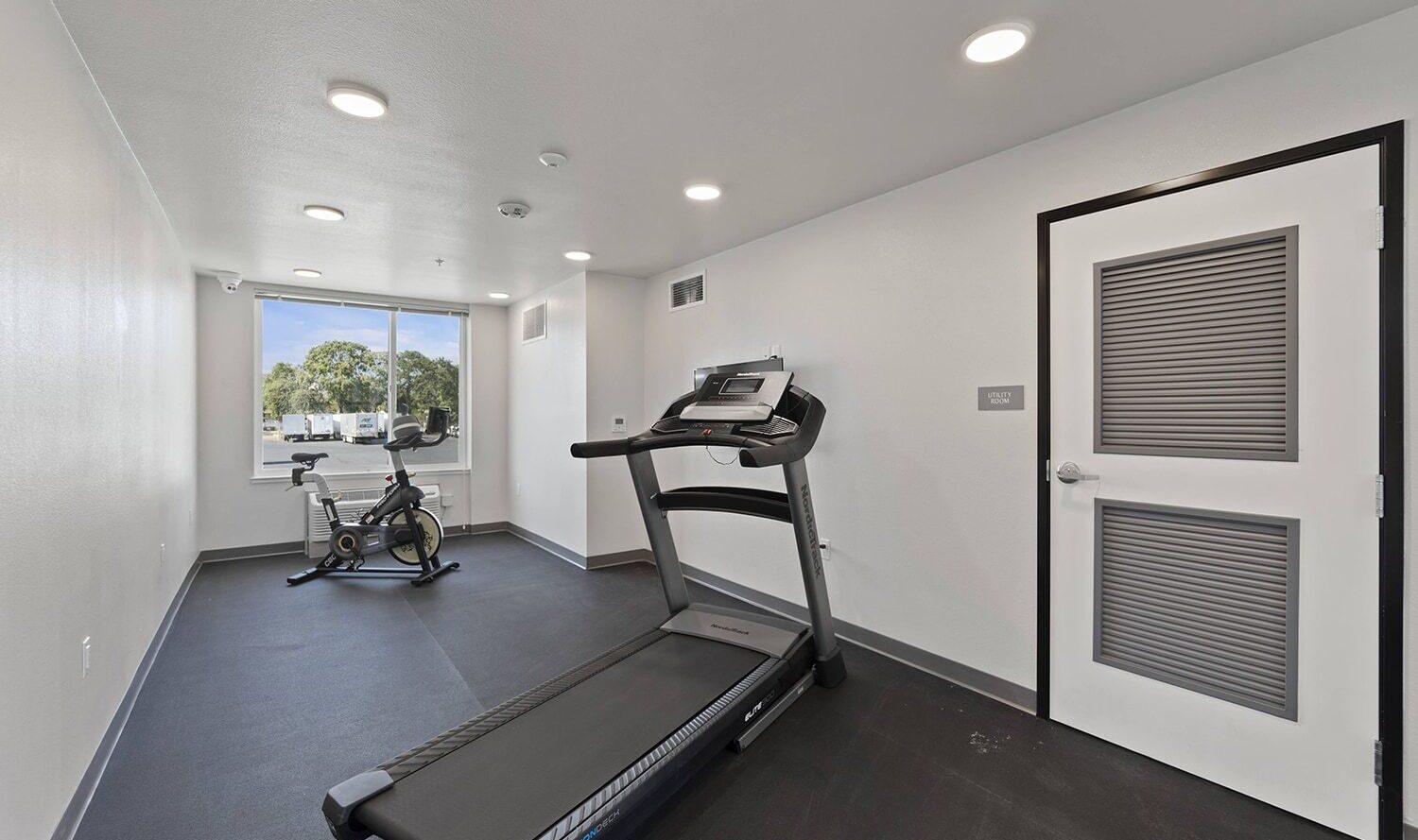 A small gym room with white walls features a treadmill and a stationary bike. There is a large window on one side, allowing natural light to illuminate the space. The black floor contrasts with the bright interior.
