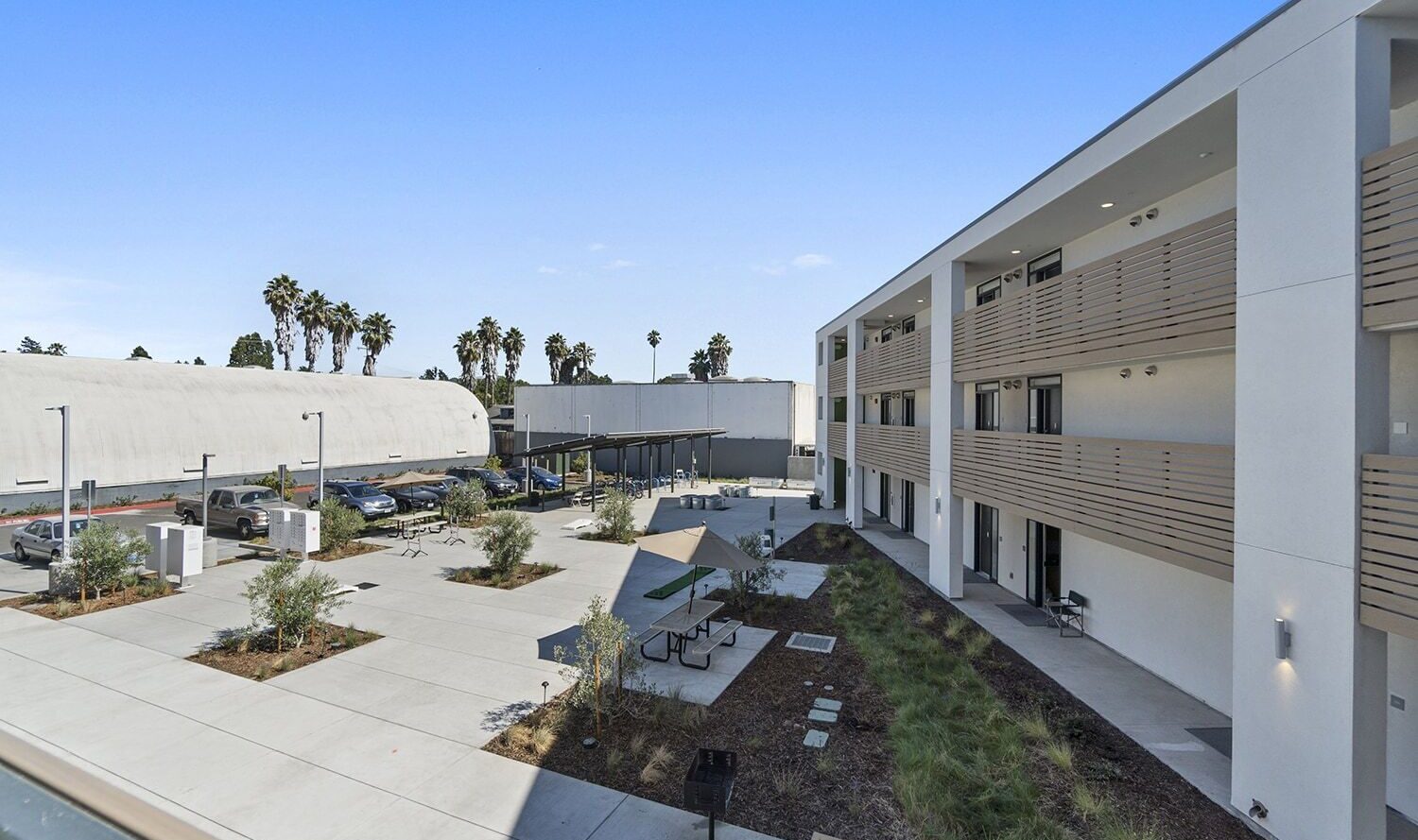 A modern apartment complex with a white exterior, featuring multiple balconies. The courtyard includes a seating area with umbrellas, landscaped greenery, and a parking lot with several cars. In the background, palm trees are visible under a clear blue sky.