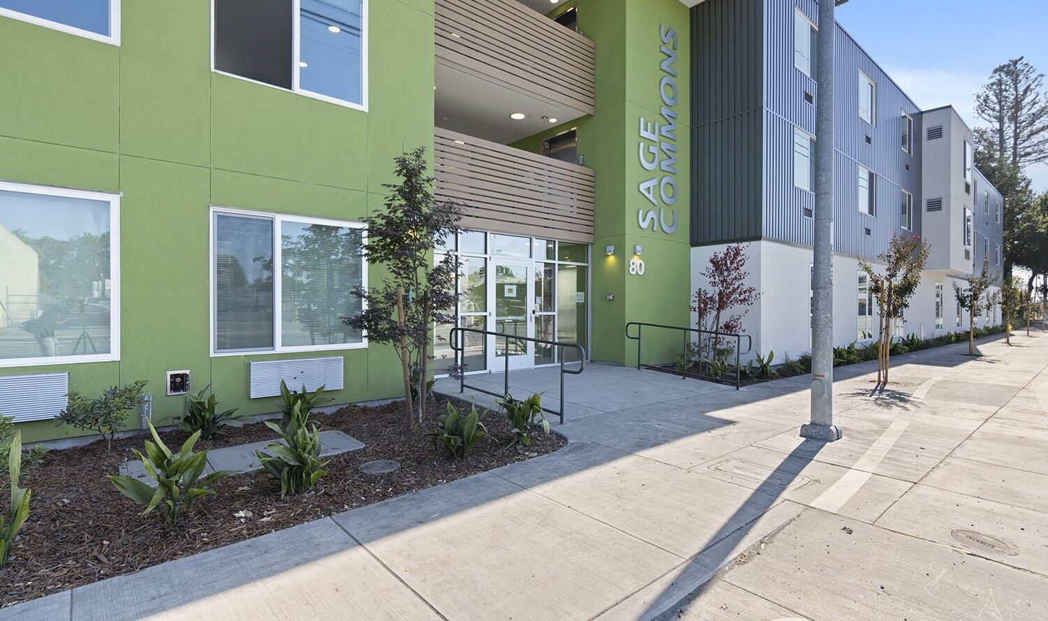 A modern apartment building with a green facade labeled Sage Commons. It features large windows, balconies, and a landscaped entrance with small trees and plants along the sidewalk.