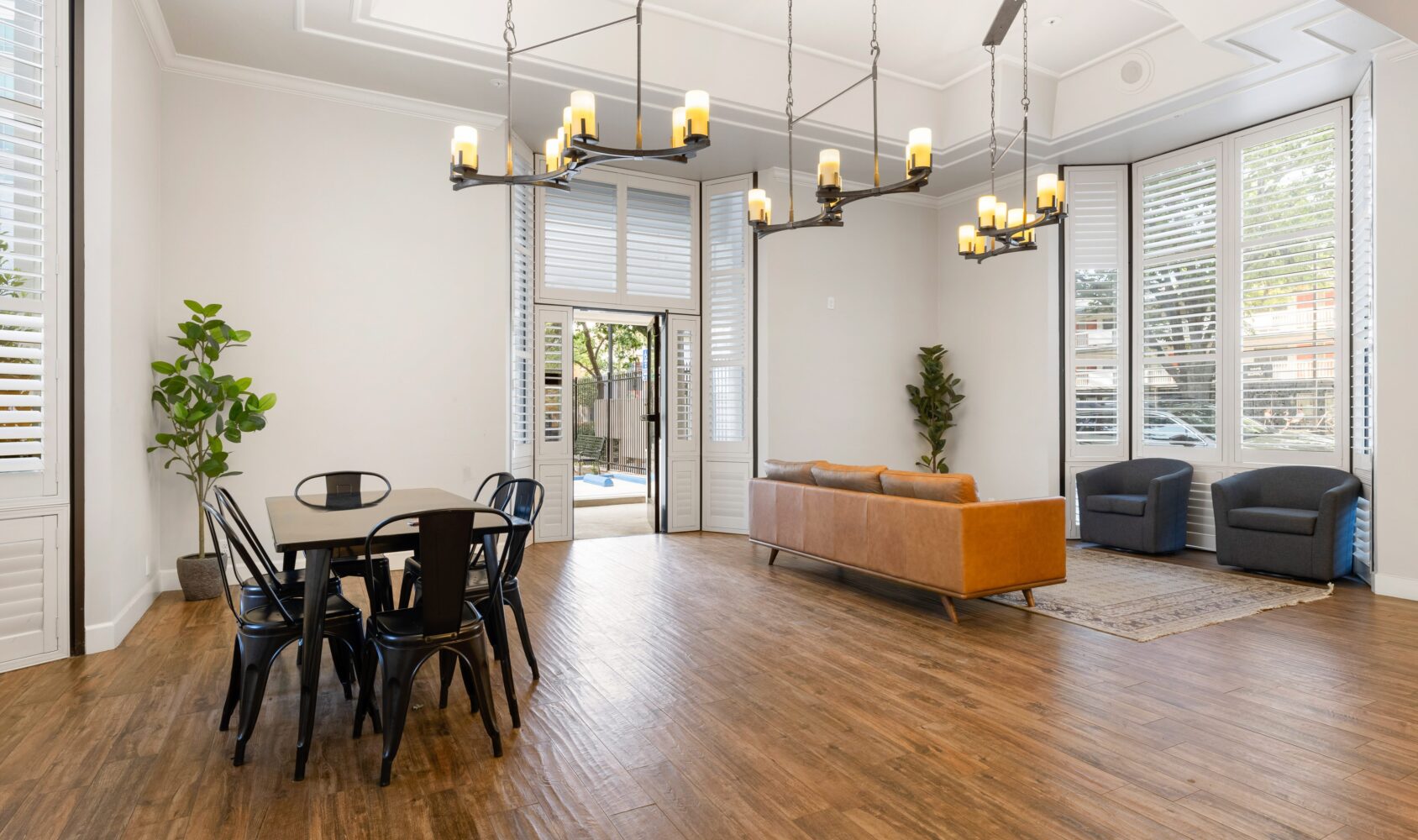 A modern living room with wooden floors, featuring a black dining table with chairs, a brown leather sofa, two gray armchairs, and large windows with white shutters. Two contemporary chandeliers hang from the ceiling, and greenery adds a touch of nature.