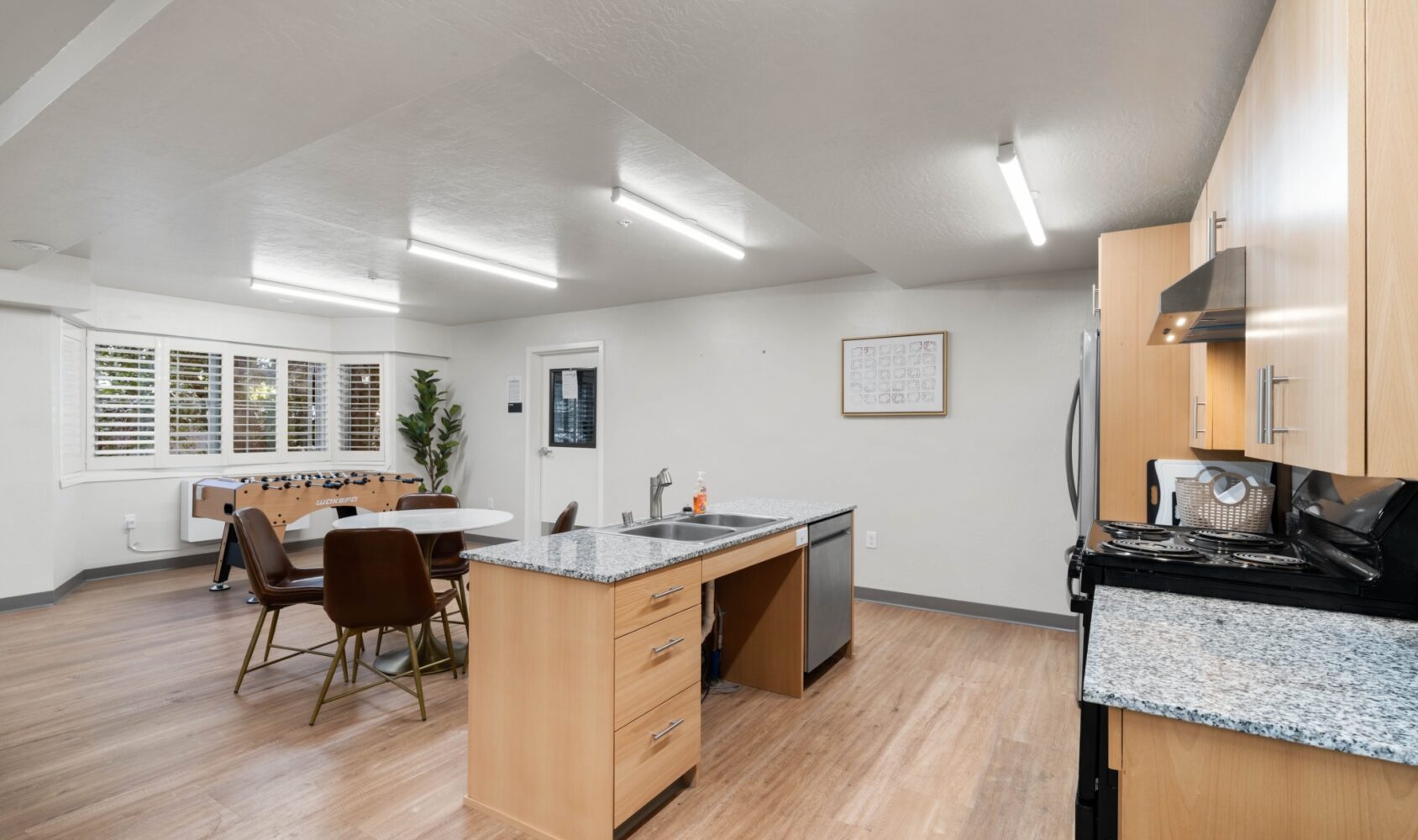 A modern kitchen and dining area featuring light wood flooring and cabinets. A circular dining table with brown chairs is in the background, alongside a foosball table and a potted plant. Overhead lighting brightens the space.