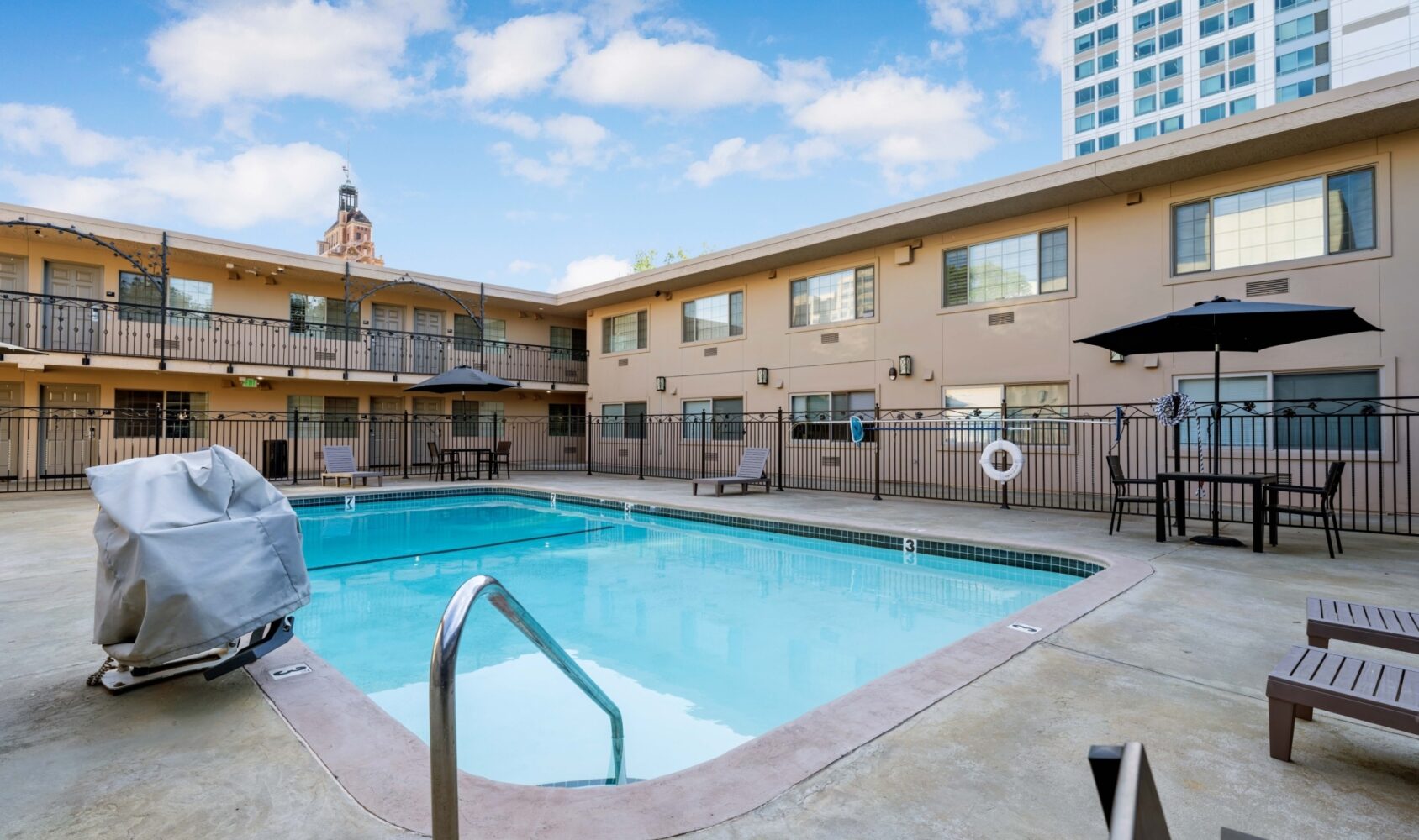 Outdoor swimming pool area within an apartment complex, surrounded by a black fence and lounge chairs. Two-story beige building in the background, with a cloudy sky above. One pool chair is covered, and a tall building is visible in the distance.