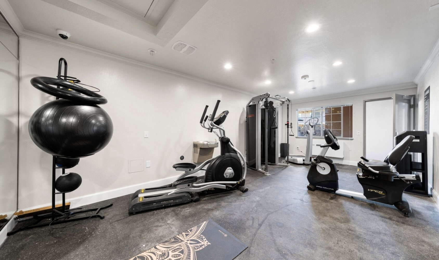 A compact gym room with sleek exercise equipment, including an elliptical, stationary bike, weight machine, and resistance balls. The room has a large mirror, white walls, and a gray floor. The lighting is bright and even.