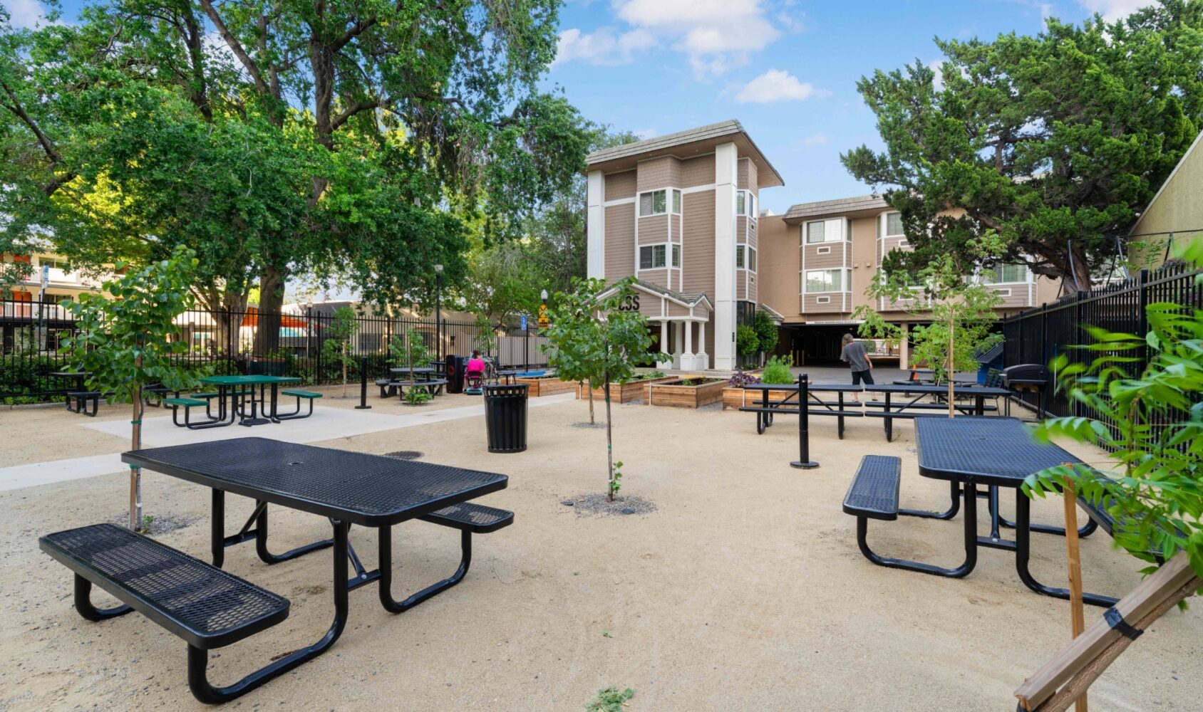 An outdoor courtyard with several black picnic tables and benches on a sandy surface. A few small green trees are scattered around, and a multi-story building is in the background. A person in a pink shirt walks nearby.