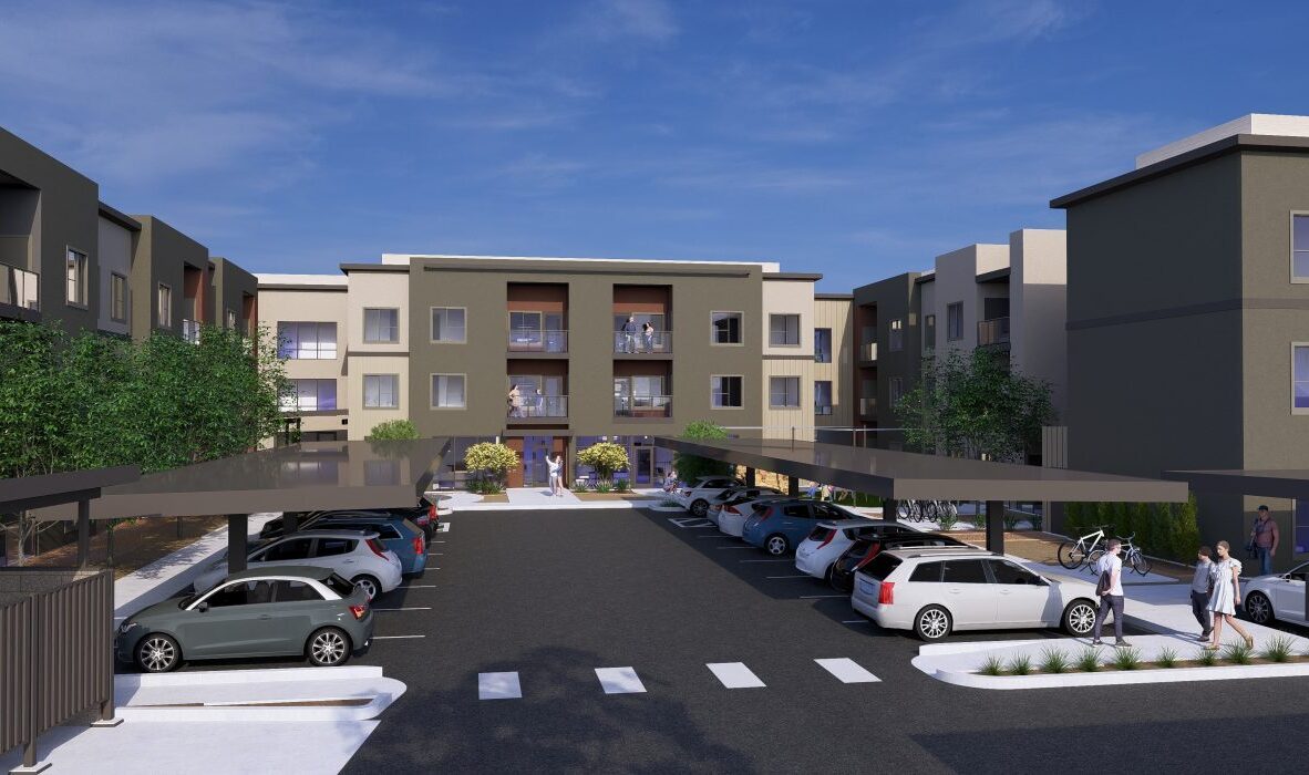 A modern apartment complex with three-story buildings, surrounded by parked cars under covered parking. The entrance features a path leading through landscaped greenery. The sky is clear and blue.