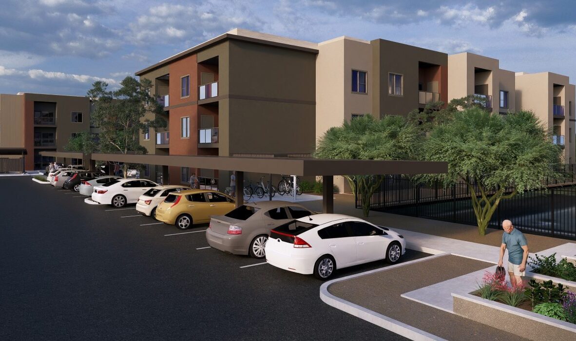 A modern apartment complex with multiple floors, featuring a parking lot with covered spaces and several cars. A man walks along a landscaped path near the entrance, and bicycles are parked under the shelter. Trees and shrubs are present.