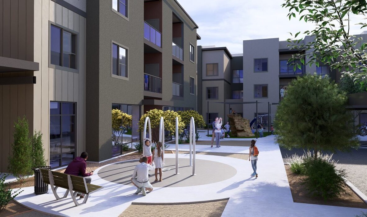 A modern apartment complex with several people gathered around a circular water fountain in the courtyard. The building is three stories high with balconies. There are green shrubs and trees, along with benches and a small bicycle rack.