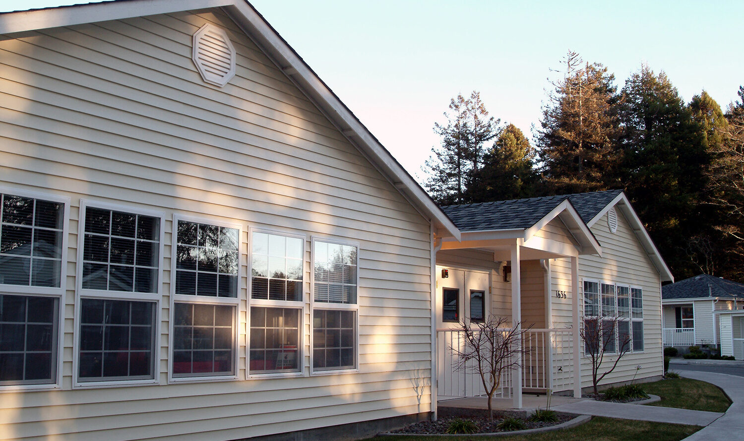 A single-story house with light yellow siding and multiple large windows. The front entrance has a small porch with a railing. The property is surrounded by trees, and the sky is clear.