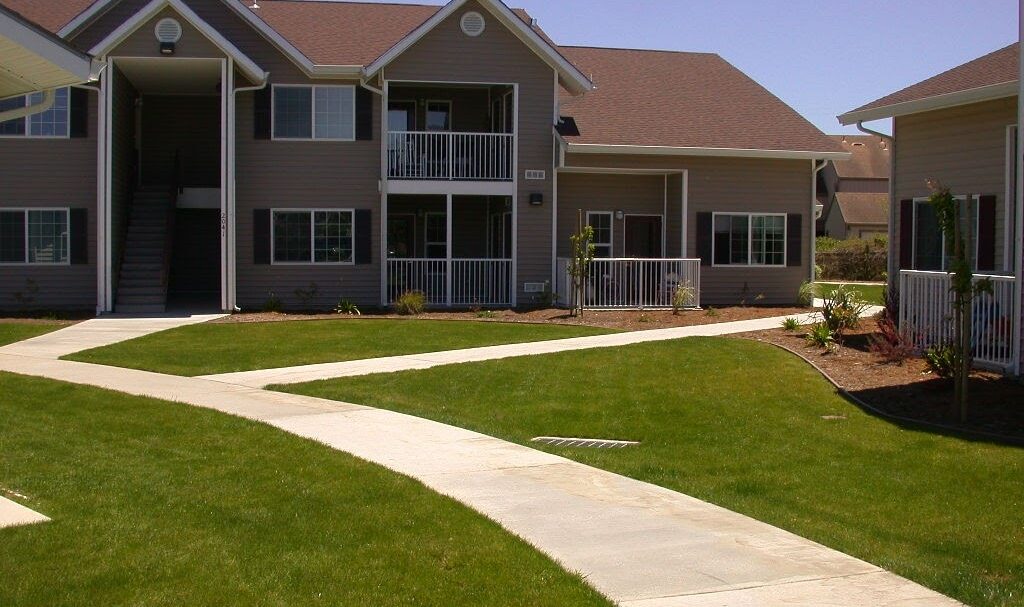 A two-story apartment building with beige siding and white trim. There are stairs leading to the second floor, balconies, and well-maintained lawns with winding concrete walkways. The sky is clear and blue.