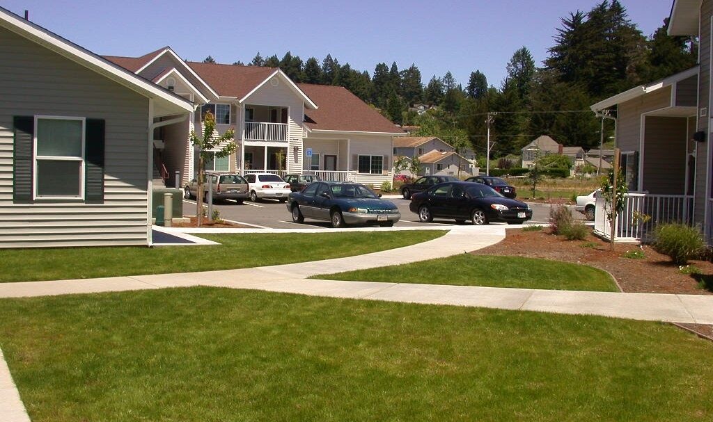 A suburban neighborhood with several houses, manicured lawns, and a few parked cars in a driveway. The sky is clear and trees can be seen in the background. Paths intersect at various points on the grass.