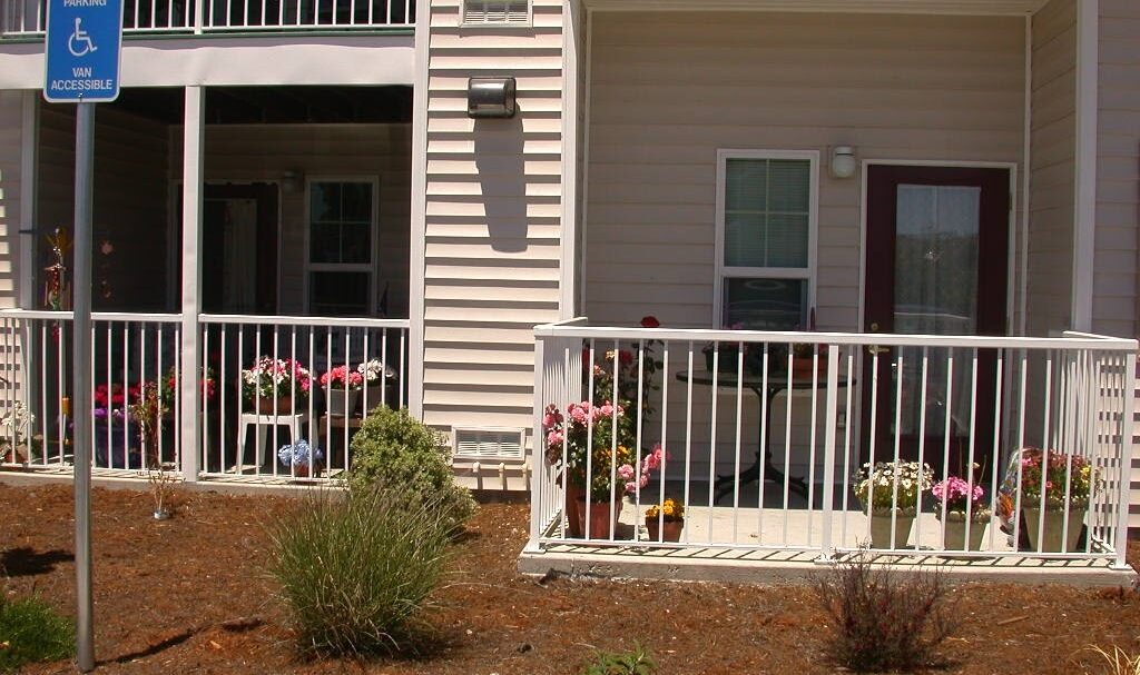 A ground floor apartment with a small porch, surrounded by a white railing. Potted plants and flowers decorate the porch. A sign indicates reserved parking for vans, and the area is landscaped with mulch and shrubbery.
