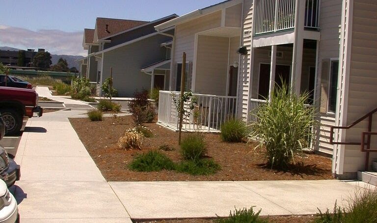A row of modern, two-story townhouses with beige siding and balconies. The buildings are surrounded by landscaped gardens and a paved walkway. The sky is clear and sunny. Cars are partially visible to the left.