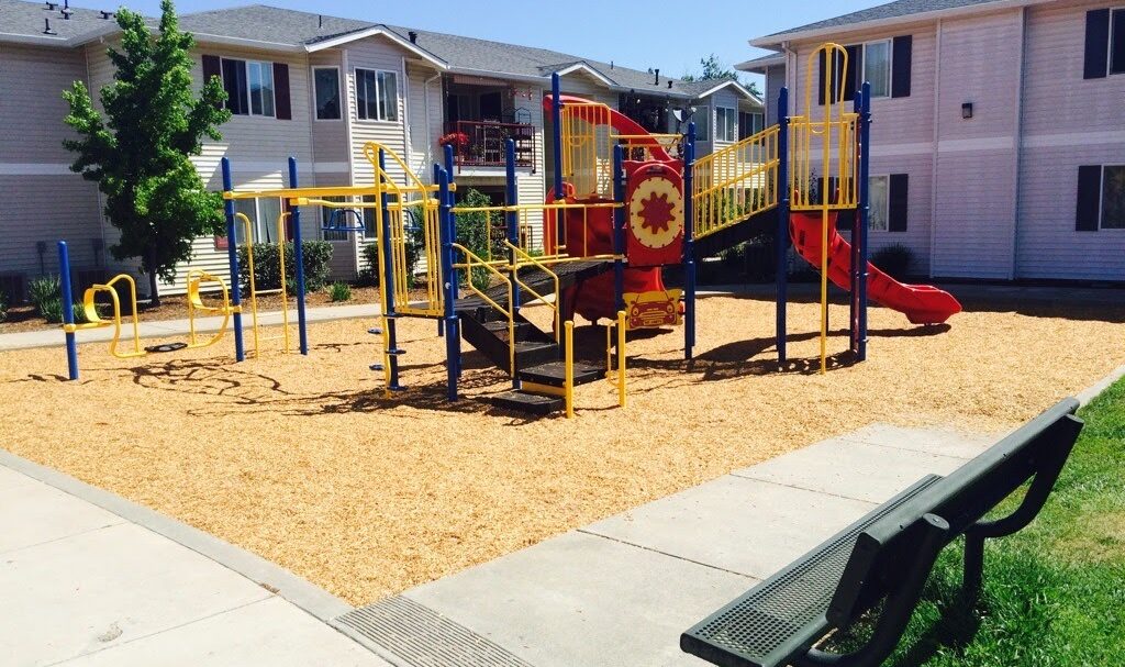 A playground with red and yellow equipment, including a slide and climbing structures, is surrounded by wood chips. Its situated in a courtyard between residential buildings, with a bench and green grass nearby.