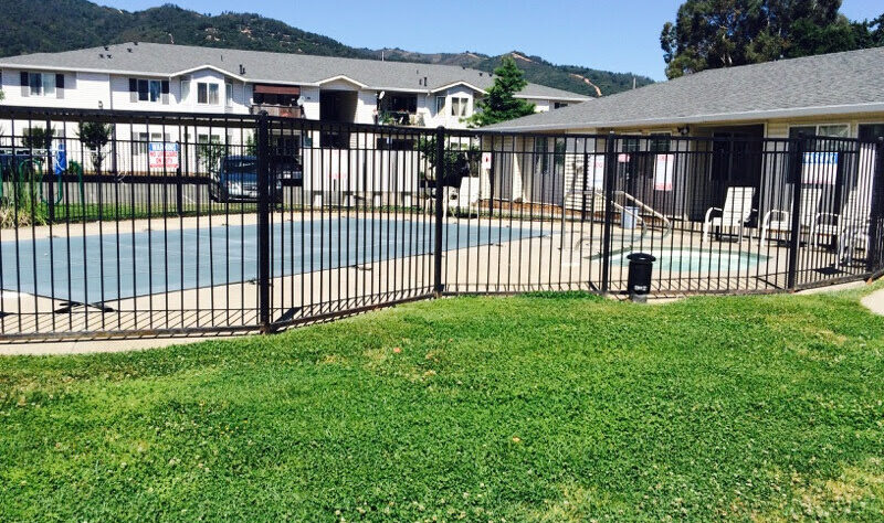 A fenced outdoor pool with surrounding concrete deck is adjacent to residential buildings. The area features lounge chairs and green grass in the foreground. Hills and trees are visible in the background under a clear blue sky.