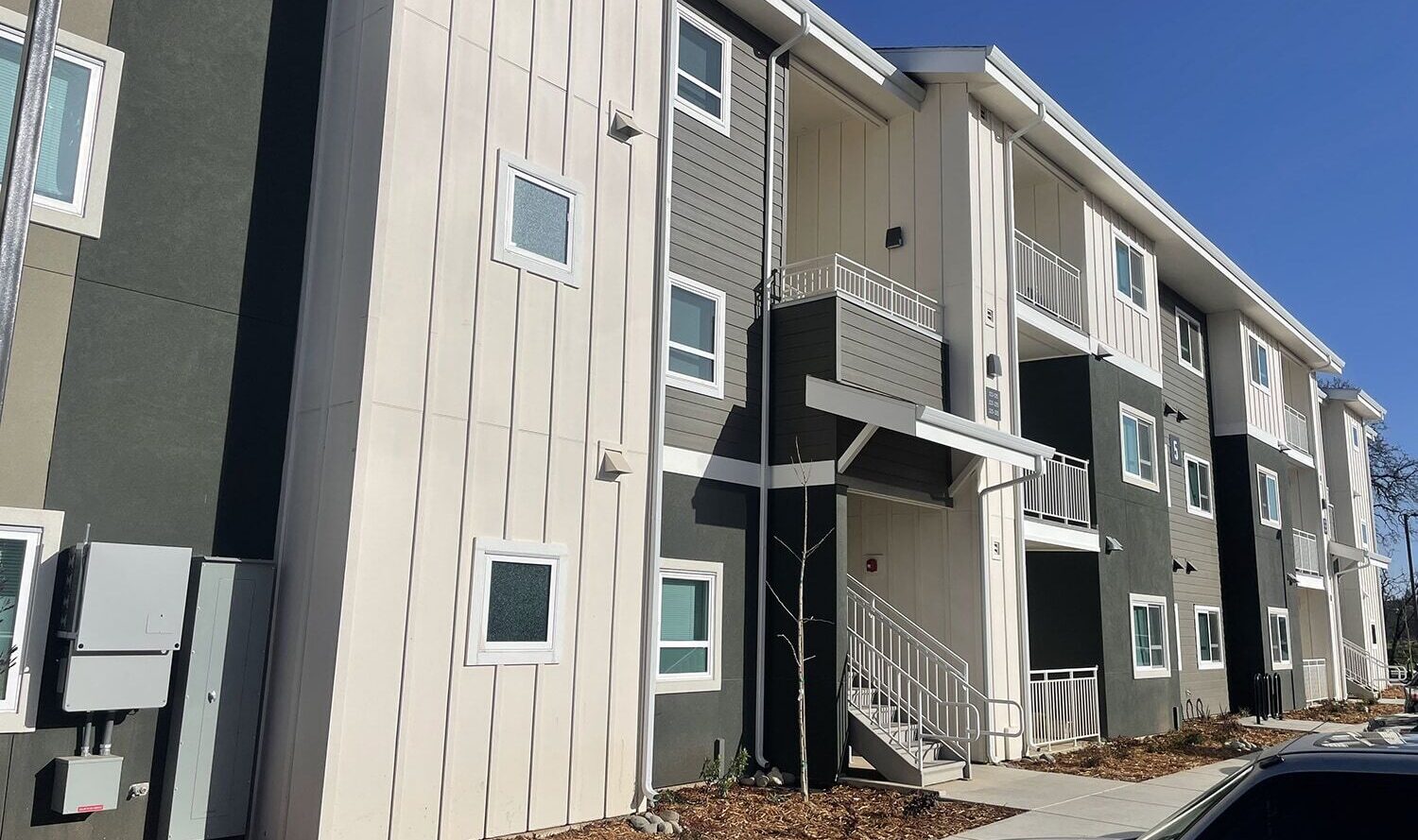 A modern three-story apartment building with a mix of green and white panels. The entrance has a staircase leading to upper floors. The sky is clear and blue, and there is landscaping with mulch and plants near the sidewalk.