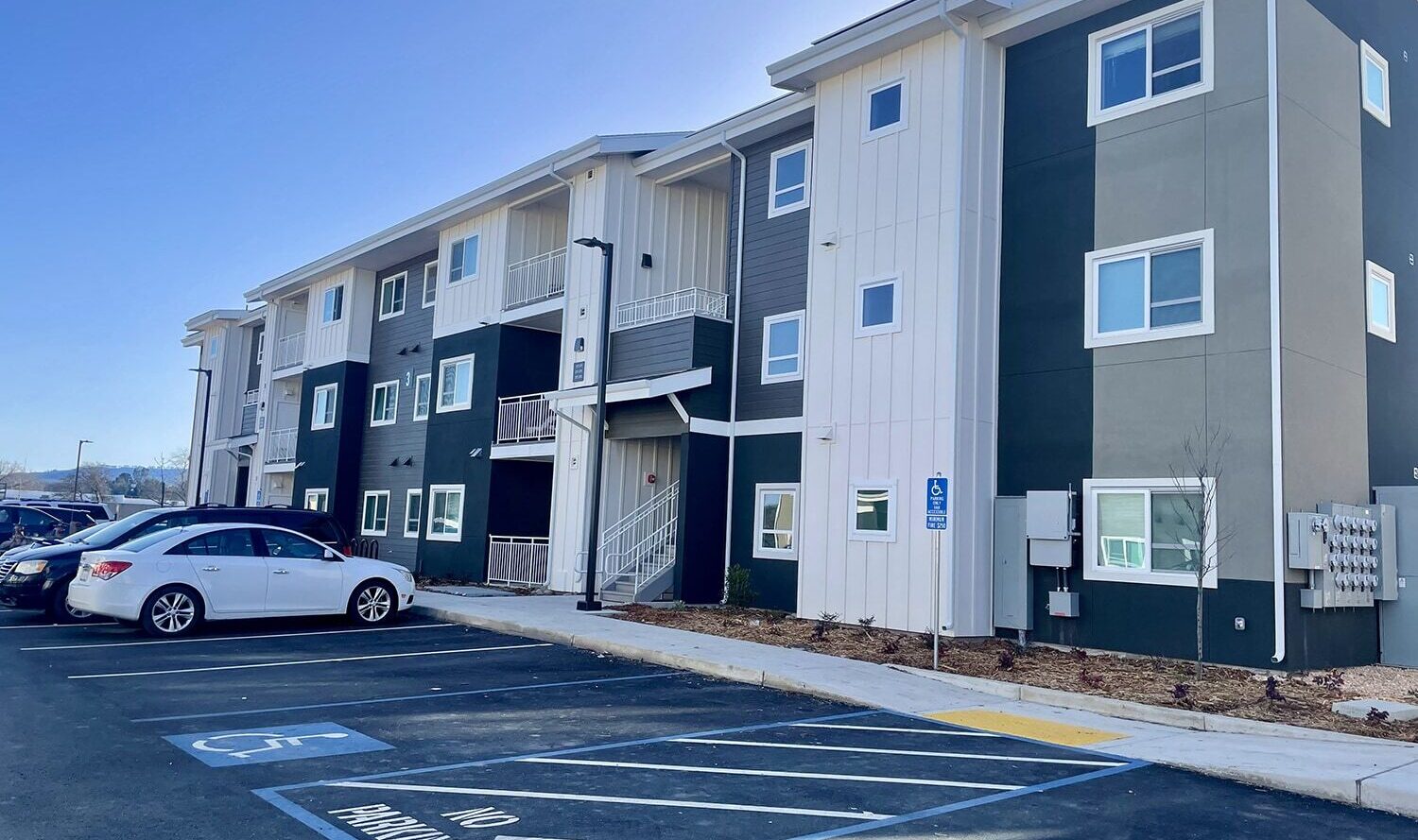 A modern three-story apartment complex with a mix of dark and light gray siding. The parking lot in front has designated spaces, including a handicap spot. The sky is clear and blue.