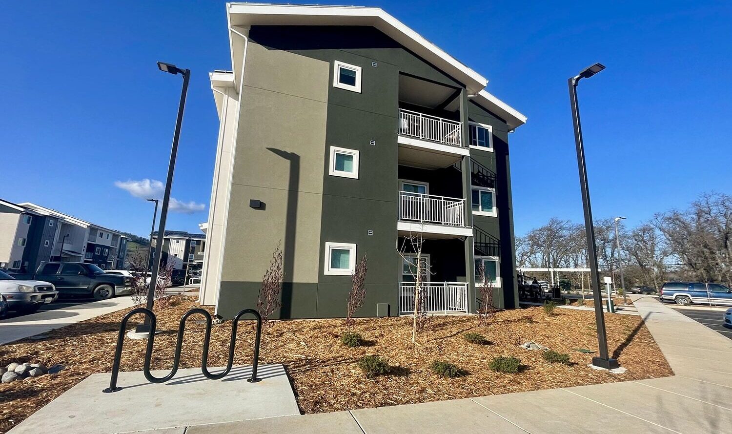 Three-story apartment building with balconies, surrounded by landscaped areas. Bike rack and parking spaces are visible in the foreground. The sky is clear and blue.