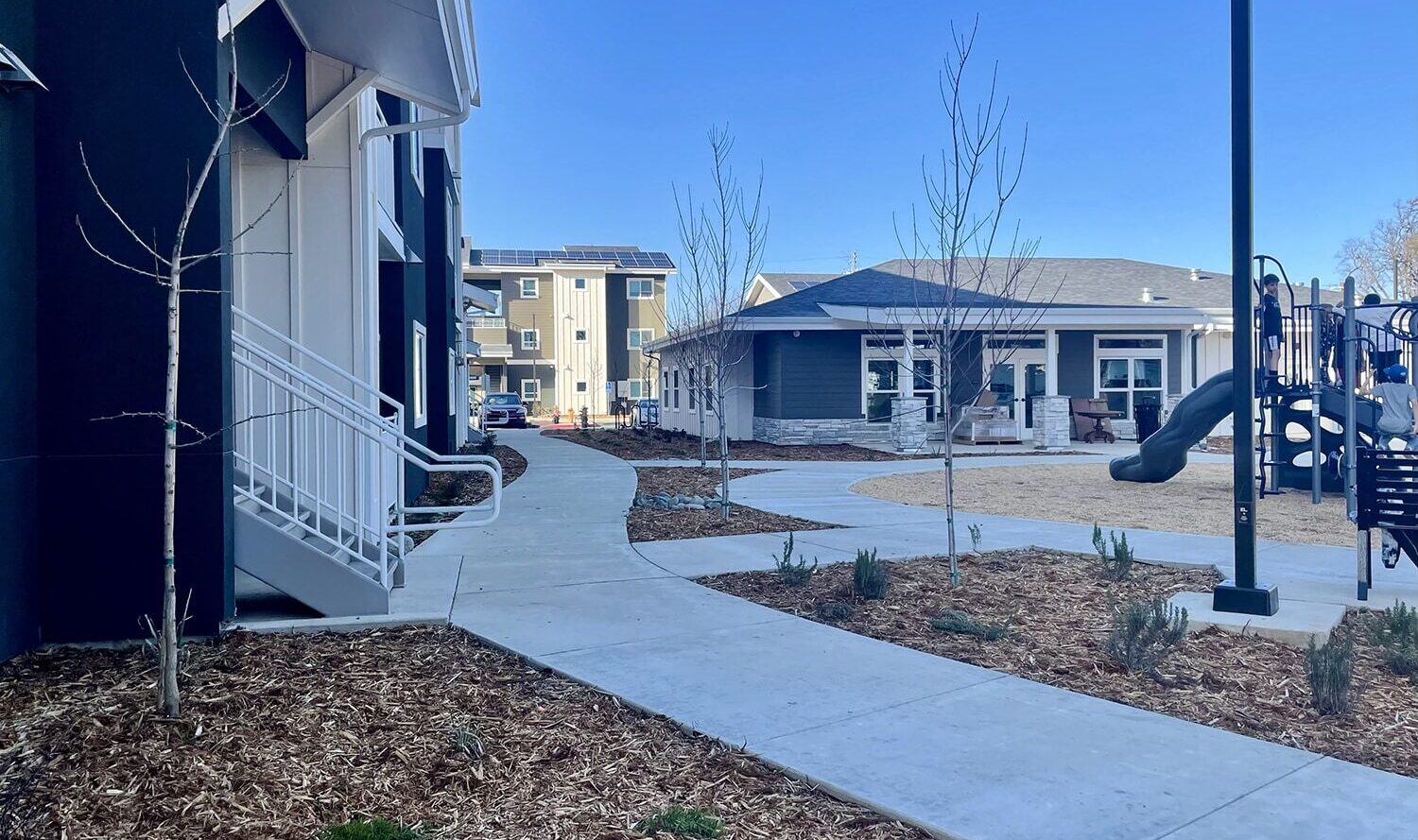 A modern apartment complex with a landscaped walkway leading to a playground. The area features young trees, mulch landscaping, and a clear blue sky. Buildings surround the play area, which includes a slide and climbing structures.