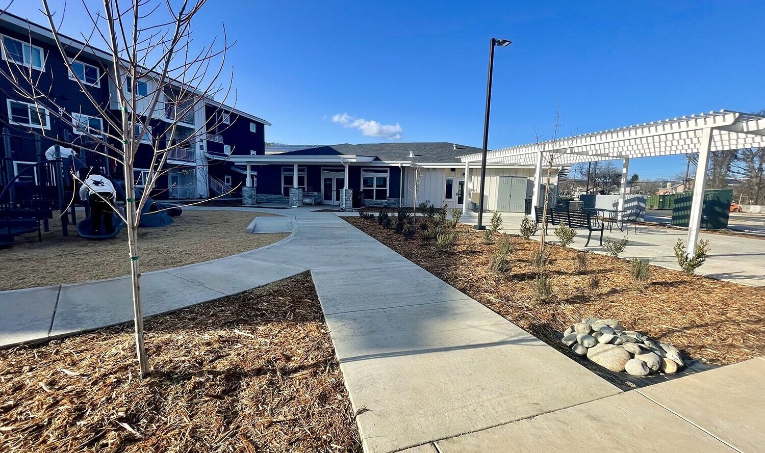 A modern building with white panels and large windows stands under a clear blue sky. In the foreground, a paved pathway winds through a landscaped area with young trees and mulch, leading to an outdoor seating area with a white trellis.